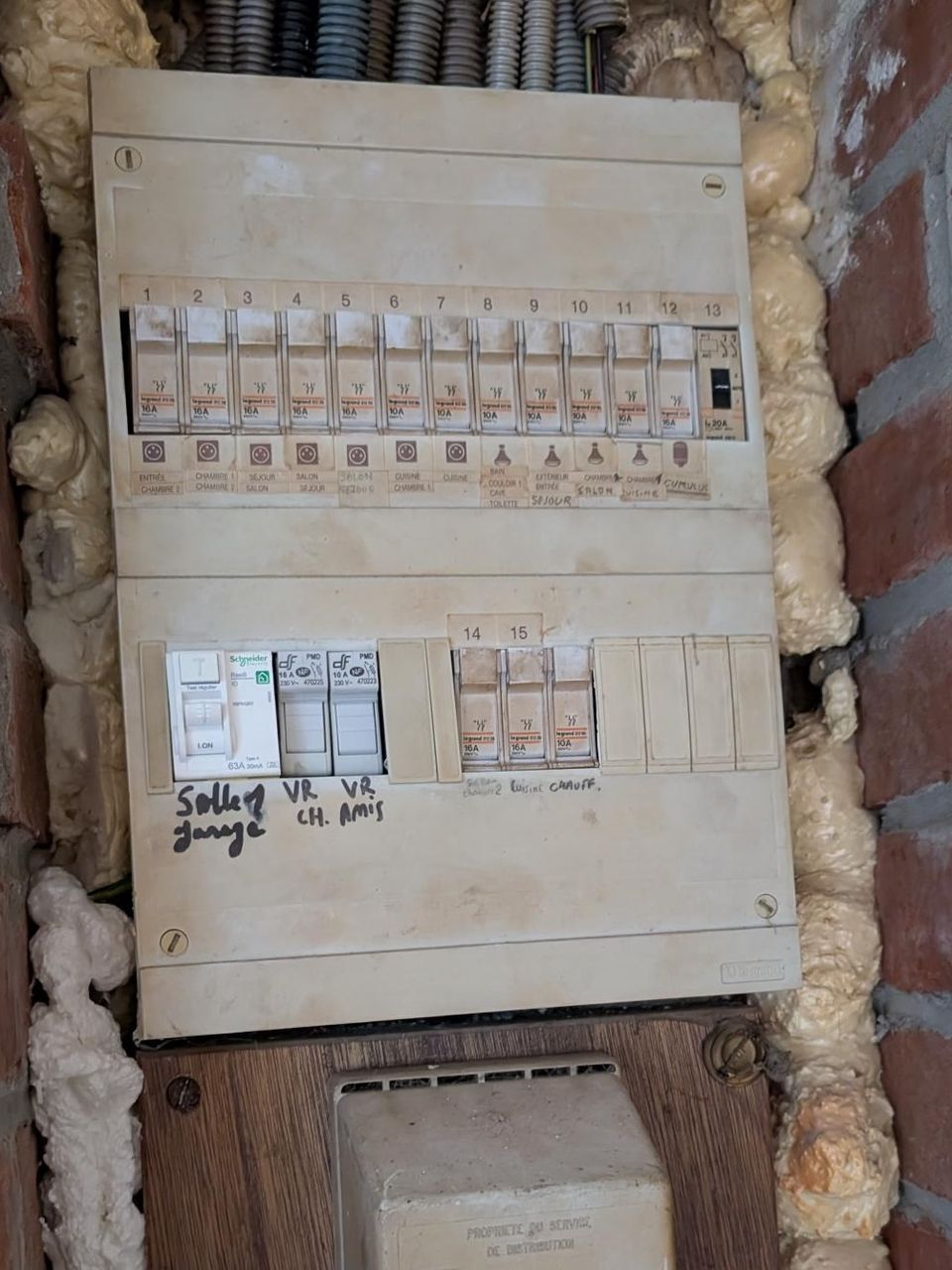 Vue rapprochée d'un tableau électrique d'origine et dangereusement vétuste dans une maison ancienne à Loos-en-Gohelle. Il est composé de vieux fusibles à vis et de deux disjoncteurs Schneider non protégés par des différentiels, le tout entouré de mousse PU expansée et surmonté d'un enchevêtrement de câbles.