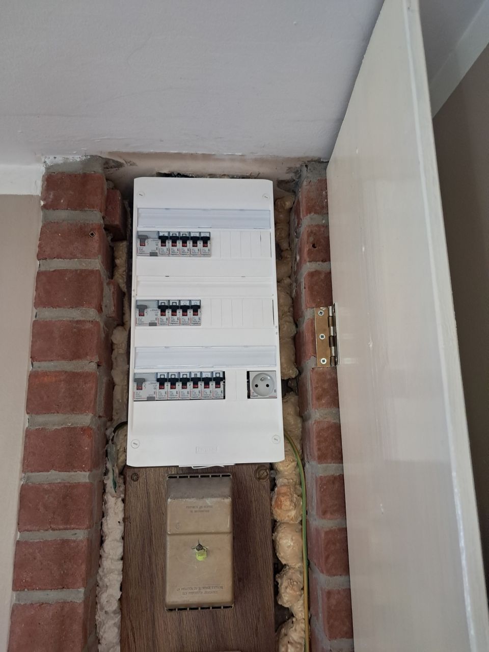 Vue nette d'un tableau électrique principal Legrand entièrement mis aux normes et sécurisé, installé dans un renfoncement bordé de briques rouges apparentes dans une maison ancienne à Loos-en-Gohelle. Le tableau blanc et moderne, équipé de disjoncteurs différentiels et d'une prise intégrée, contraste avec une vieille boîte de distribution visible au-dessous.